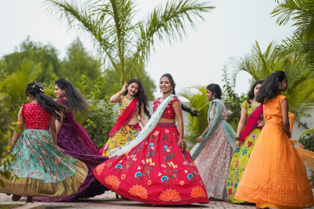 Group of South Asian women wearing vibrant traditional saris dancing joyfully outdoors.
